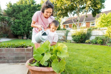 Mother and son watering vegetables in pots garden in Backyard on Sunny Summer Day. Boy helps mom take care of kitchen garden, woman teaches son to take care of plants. Time together. Active childhood.