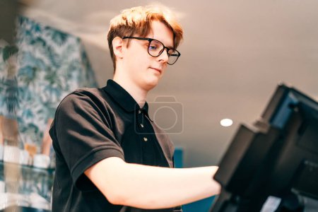 Young waiter serving customer at cash point in cafe. Man working with POS terminal. Cashier, barista checking for payment receipt. Hospitality, server and preparing a slip at the till in coffee shop.
