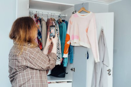 Woman taking photo of the sweater using her smartphone for selling or donating her clothes. Decluttering , Sorting Clothes, And Cleaning Up wardrobe. Reuse, second-hand concept. Conscious consumer