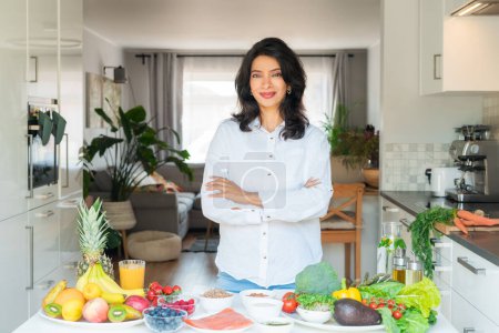 Confident smiling middle aged female Indian Nutritionist expert arms crossed in front of kitchen table full of fruits and vegetables. Healthy diet habits and wellness lifestyle, weight loss program