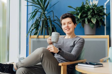 Smiling Neutral gender middle aged business woman in casual clothing relaxing with coffee cup on the armchair. Female professional enjoying coffee drink in office lounge. Take a break for recovery.