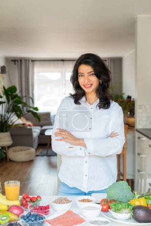 Confident smiling middle aged female Indian Nutritionist expert arms crossed in front of kitchen table full of fruits and vegetables. Healthy diet habits and wellness lifestyle, weight loss program