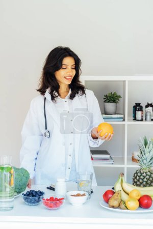 Confident smiling middle aged female Indian Nutritionist doctor holding orange in front of office desk full of fruits and vegetables. Healthy diet habits and wellness lifestyle, weight loss program