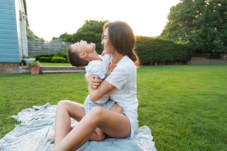 Téléchargez les photos : Joyeux garçon d'âge préscolaire mignon s'amuser avec sa mère, embrasser, rire sur l'herbe verte. Jeune maman et enfant étreignant avec amour, affection, tendresse sur le fond de pelouse arrière-cour sur le coucher du soleil - en image libre de droit
