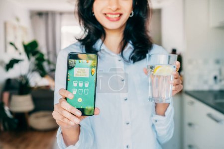 Healthy habit to drink water. Blurred Indian woman with glass of pure water with lemon holding phone with active mobile app to control body hydration, Tracking water balance. Selective focus