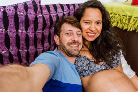 Foto de Retrato selfie pareja latina, hombre adulto caucásico con barba y ojos azules con su esposa, mujer adulta brasileña embarazada sonriendo, feliz sentado en el sofá - Imagen libre de derechos