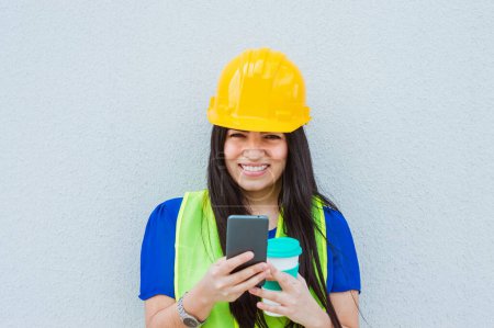Porträt einer Ingenieurin mit Schutzhelm und reflektierender Weste, die mit einer Wand im Hintergrund auf der Straße steht, lächelt, ihr Handy, eine Tasse Kaffee in die Kamera hält