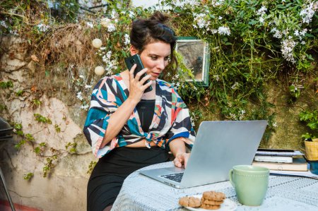 Foto de Caucasian adult woman of French ethnicity, working on the terrace with her laptop and listening to a voice memo from her phone, with a cup of coffee and cookies on the table, copy space. - Imagen libre de derechos