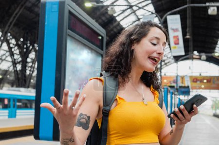 Téléchargez les photos : Young latin traveler woman of argentinian ethnicity arguing by video call with her boyfriend on the phone while waiting for the train standing on the platform. - en image libre de droit