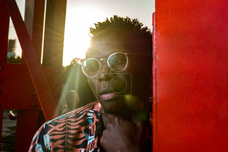 Téléchargez les photos : Portrait coucher de soleil d'un jeune homme africain, avec des lunettes, des cheveux courts et des vêtements d'été, est sérieux et réfléchi en regardant la caméra, appuyé contre des poutres rouges dans un parc public, gros plan. - en image libre de droit
