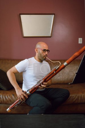 Photo for Young hispanic latino man of venezuelan ethnicity, bald and wearing glasses. at home sitting on a leather sofa, relaxed playing the bassoon, vertical image, copy space, music concept. - Royalty Free Image