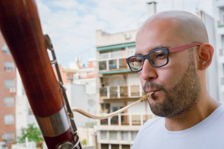 Photo for Young latino venezuelan saxophonist man, with glasses, beard, white clothes and bald, very concentrated playing the bassoon outdoors, practicing and studying classical music. horizontal portrait. - Royalty Free Image