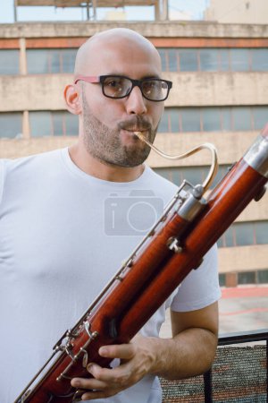 Photo for Waist-up portrait of young latino man, with beard, glasses, bald and in white clothes, saxophonist playing a bassoon standing on the balcony of his apartment in Argentina, looking at the camera. - Royalty Free Image