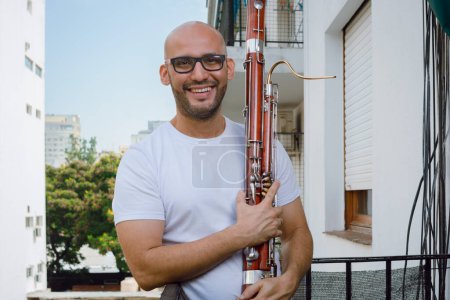 Téléchargez les photos : Portrait d'un jeune latino barbu, chauve et vêtu de blanc, musicien saxophoniste tenant un basson debout sur le balcon de son appartement regardant la caméra et souriant. - en image libre de droit