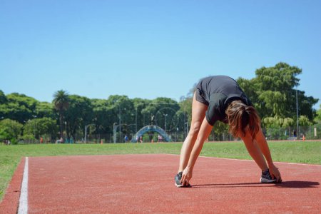 Foto de Joven latina de etnia argentina estirándose en la pista de atletismo, de pie e inclinada hacia adelante tocándose los pies con las manos, entrenando preparándose para correr. - Imagen libre de derechos
