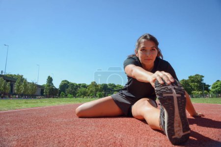 Wide angle front view of sporty young latina woman of Argentinian ethnicity, stretching and training on the running track, preparing to run and start her routine training, sport concept, copy space.