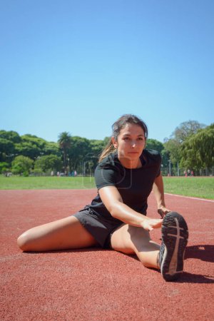 Foto de Imagen vertical de una joven latina, estirando la pierna y la cadera tocando el dedo del pie con la mano, calentándose en la pista de atletismo, vista frontal, espacio para copiar. - Imagen libre de derechos