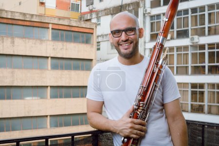 Téléchargez les photos : Portrait taille haute du jeune musicien d'orchestre vénézuélien latin barbe, lunettes et vêtements blancs, debout souriant tenant un basson sur le balcon de son appartement, regardant la caméra. - en image libre de droit