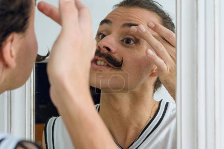 Téléchargez les photos : Reflet dans le miroir d'une jeune latino non binaire de genre se tenant à l'intérieur de sa chambre appliquant du maquillage avec son doigt sur ses paupières. - en image libre de droit