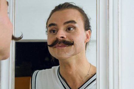 Téléchargez les photos : Réflexion dans le miroir d'un jeune Latino d'origine argentine et de sexe non binaire, moustachu, heureux de regarder fièrement après s'être maquillé. - en image libre de droit
