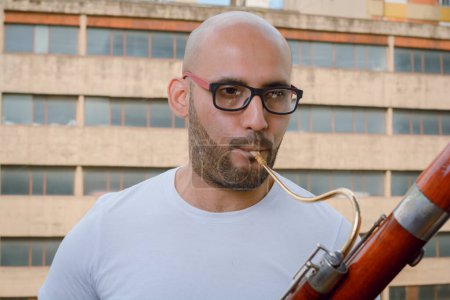 Photo for Closeup portrait of young latino man with glasses, beard and bald, wearing white t-shirt, standing playing bassoon on apartment balcony with urban buildings in background. - Royalty Free Image
