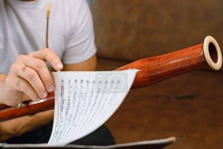 Photo for Closeup of bassoonist male hand turning sheet music page on music stand, focus on hand, with blur person on background indoors. Music concept, copy space. - Royalty Free Image