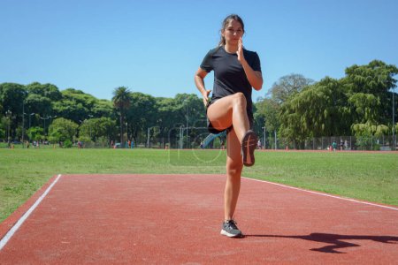 Photo for Young latina woman of argentinian ethnicity in black sportswear starting warm up exercises on the running track with one leg raised and slightly bent to the front - Royalty Free Image