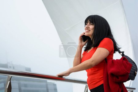 Foto de Joven latina de etnia venezolana, enamorada hablando por teléfono al aire libre, sonriendo y mirando hacia arriba con suerte, de pie en Puente De La Mujer en Buenos Aires, espacio para copiar. - Imagen libre de derechos