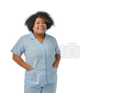 Foto de Retrato joven doctora afro-latina de etnia venezolana sonriendo y mirando a la cámara, vistiendo un uniforme azul está con las manos en los bolsillos de su bata, fondo blanco con espacio para copiar - Imagen libre de derechos