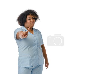 Foto de Retrato de una joven doctora afrolatina en uniforme azul, diciendo no con el dedo delante con gesto de negación en la cara, fondo blanco con espacio para copiar - Imagen libre de derechos