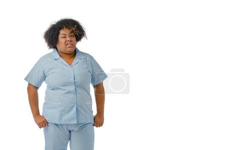 Foto de Joven enfermera afro-latina venezolana, en uniforme azul de pie mirando a la cámara con una cara asquerosa torciendo su boca, fondo blanco - espacio para copiar. - Imagen libre de derechos