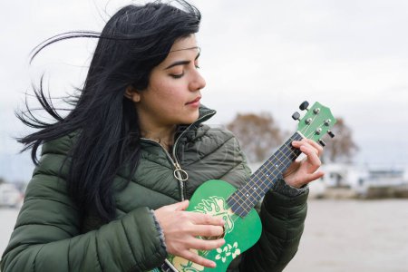 Photo for Young venezuelan latin woman, traveling outdoors on cold and windy cloudy day, playing ukulele with river in background, copy space. - Royalty Free Image