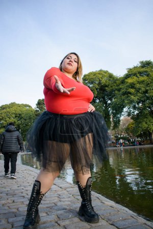 Téléchargez les photos : Jeune belle plus la taille argentine latina femme debout posant en regardant la caméra dans le parc portant une jupe en tutu noir et pull rouge. personnes et concept de mode - en image libre de droit