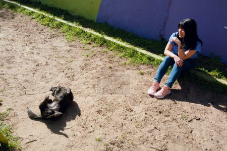 Téléchargez les photos : Vue de dessus jeune femme latine venezuelan, dans le parc assis en regardant son chien roulant sur le dos roulant sur le sol, concept de personnes et d'animaux de compagnie, espace de copie - en image libre de droit