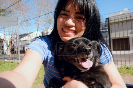 Téléchargez les photos : Selfie portrait de jeune femme latine, heureux sourire assis avec son chien dans le parc au coucher du soleil, en regardant la caméra, vue de face, mode de vie concept. - en image libre de droit