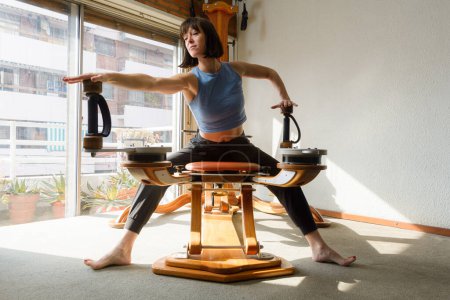 Front view of Argentinian Latina woman, with short hair and blue top, sitting stretching and working her shoulders and arms with exercise machine at home in morning.