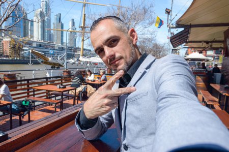 Téléchargez les photos : Homme d'affaires caucasien d'origine française, souriant joyeusement en vacances en Argentine, au restaurant à Puerto Madero assis en train de prendre des photos selfie avec son téléphone - en image libre de droit