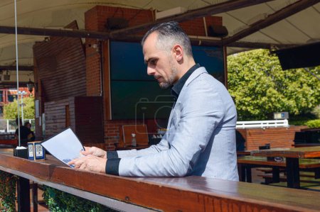 Photo for Caucasian adult business man, sitting at bar outside restaurant on sunny day in Buenos Aires, reading menu to order food - Royalty Free Image