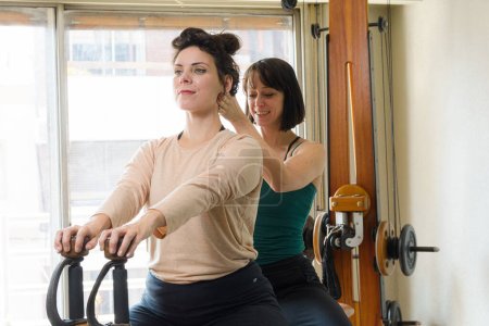 Foto de Instructora de ejercicios femenina latina adulta corrigiendo la postura espinal de su estudiante sentada en la máquina de ejercicios en casa. - Imagen libre de derechos