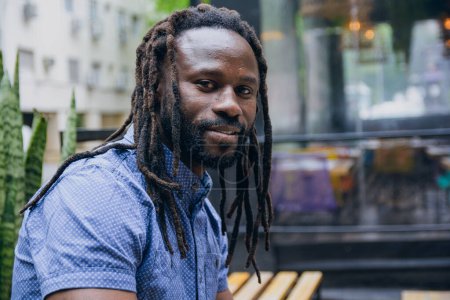 Photo for Profile view portrait of African man with beard, dreadlocks and shirt, sitting outside cafe looking at camera alone and calm, copy space. - Royalty Free Image