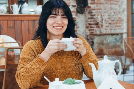 Photo for Young latin woman with black hair and brown clothes, comfortable enjoying her cup of tea, smiling looking at the camera placidly, lifestyle concept, copy space - Royalty Free Image