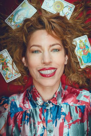 Téléchargez les photos : Portrait de belle femme heureuse lecteur de tarot souriante regardant caméra couchée sur la table rouge, avec des cartes autour de sa tête et sur ses cheveux, image verticale - en image libre de droit