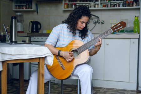 Photo for Front view of young Latin woman venezuelan, with curly hair, classical music student alone at home sitting concentrated practicing major scale on acoustic guitar - Royalty Free Image