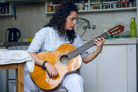 Photo for Front view of young Latin woman venezuelan, with curly hair, classical music student alone at home sitting concentrated practicing major scale on acoustic guitar - Royalty Free Image