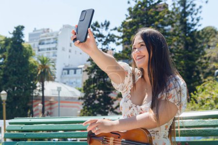young Latina busker violinist woman sitting with her violin on bench outdoors, in plaza, smiling, taking selfie photo with her phone, technology concept, copy space.