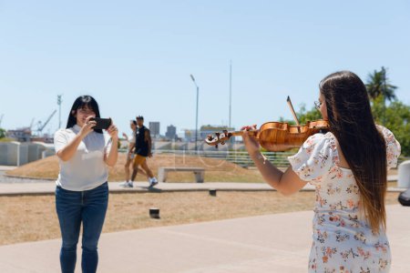 rear view of young latin woman busker violinist content creator playing violin outdoors while another girl films her for social networks.
