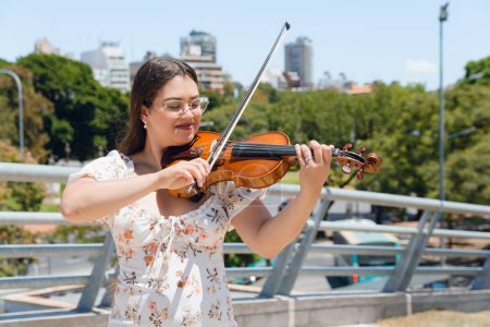 linda latina venezolana busker mujer feliz sonriendo de pie tocando el violín al aire libre al mediodía, en la ciudad buenos aires, con árboles y ciudad en el fondo, espacio de copia