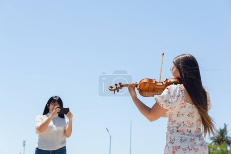 Rückansicht der jungen lateinamerikanischen Frau busker Geigerin Content Creator Geige spielen im Freien, während ein anderes Mädchen filmt sie für soziale Netzwerke.