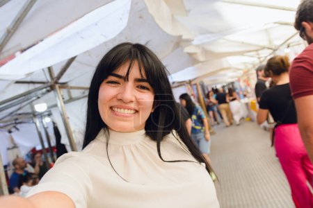 Portrait of young Latin traveler woman with long black hair and white clothes, on vacation taking selfie photo with her phone looking at camera smiling, mobile phone camera perspective, copy space.