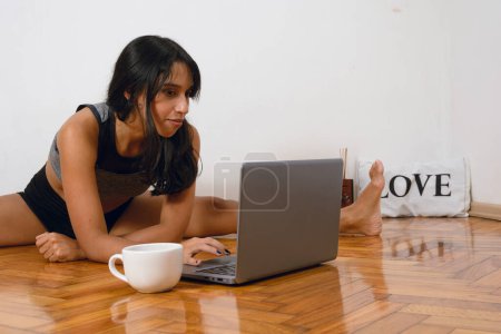 young brunette latin woman with long hair, wearing sports clothing, yoga teacher with legs open sitting on floor with laptop planning online classes, copy space.